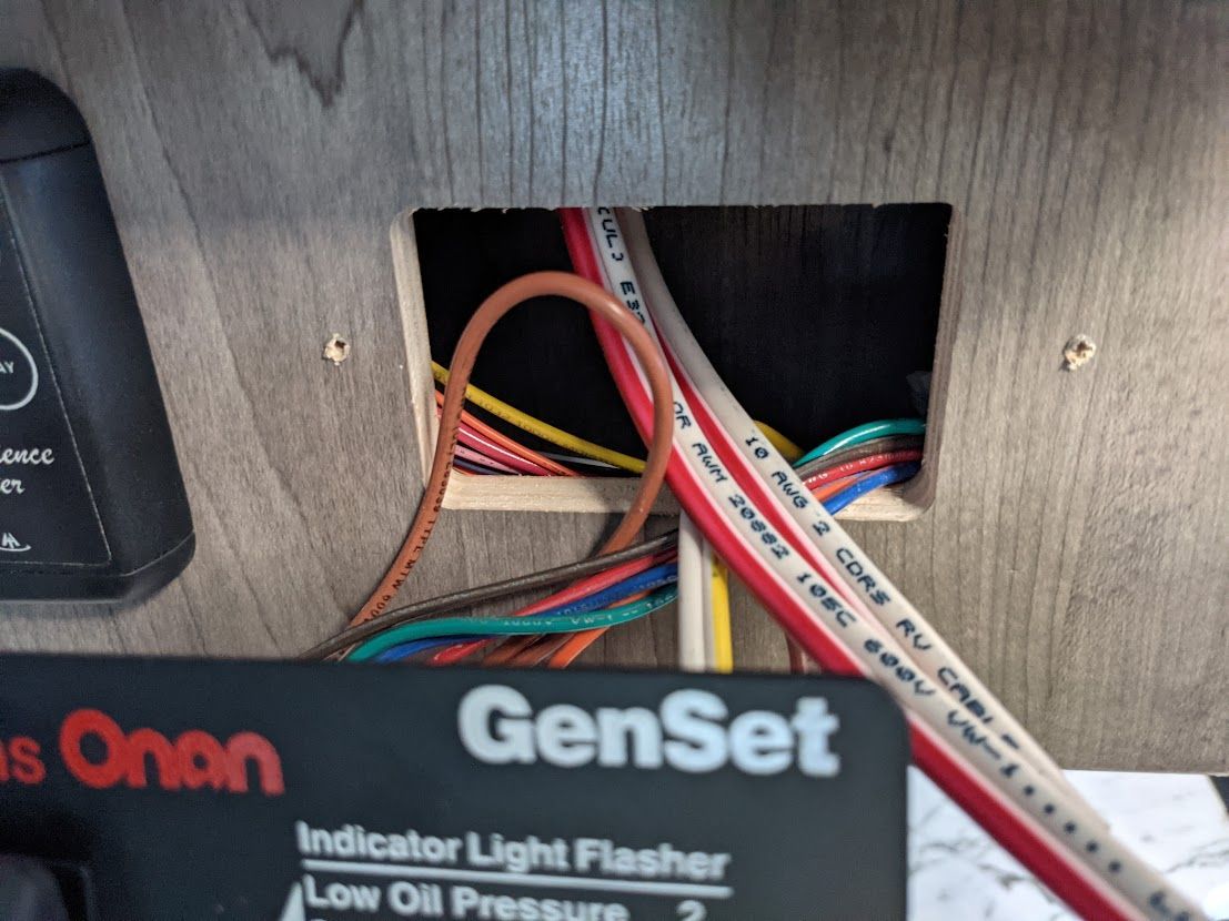 Wires through a hole in RV cabinetry, above an Onan GenSet control panel.