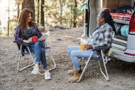 Two people sit in folding chairs beside a white van, talking at a campsite with trees in the background.