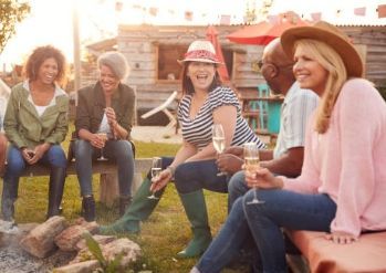 Friends sitting around a campfire outdoors, smiling and chatting in casual clothes and hats.