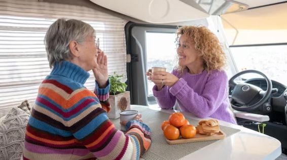 Two people share drinks at a table inside a camper van, with oranges and a warm smile.