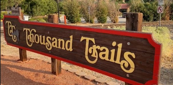 Red wooden sign reading “The Thousand Trails” with tan lettering beside a roadside trail area.