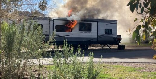 RV trailer on fire, with flames and thick smoke rising near a roadside in a wooded area