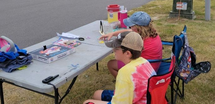 Two children sit at an outdoor folding table with snacks and crafts beside a road.