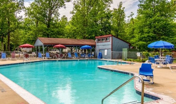 Outdoor swimming pool with blue umbrellas, lounge chairs, and a clubhouse surrounded by trees
