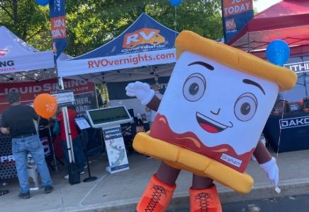 Smiling white mascot s'more in orange pants at an outdoor RV show booth with tents and balloons