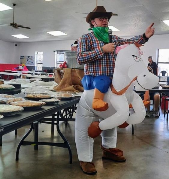 Person in a cowboy hat and mask riding a white stuffed horse beside trays of food in a bright hall