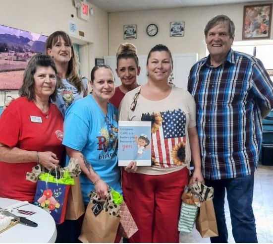 Five people posing indoors with gift bags and a certificate in a community room