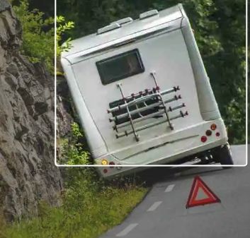 White camper van tilted on a curving mountain road beside a rock cliff and trees.