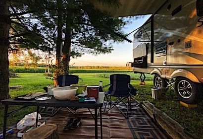 Camping table and chairs beside an RV under trees, with a grassy field and sunset in the background