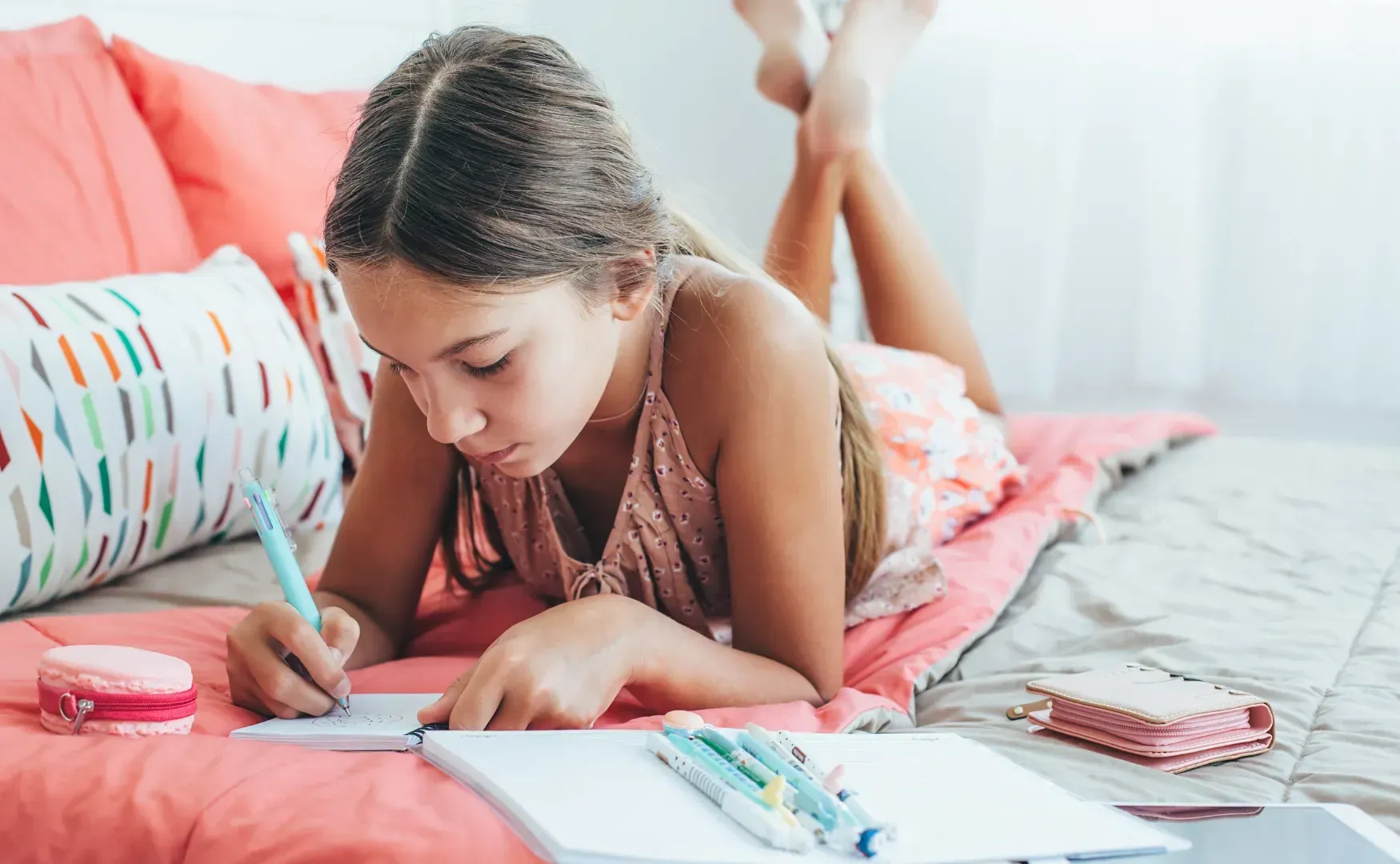 Girl lying on a bed writing in a notebook with a pen; surrounded by pillows and art supplies.