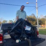 Man loading a large black bag into a truck bed outside; blue sky in background.