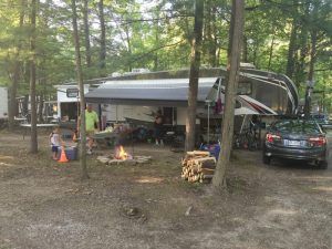 A group of people are sitting around a campfire in front of a rv in the woods.