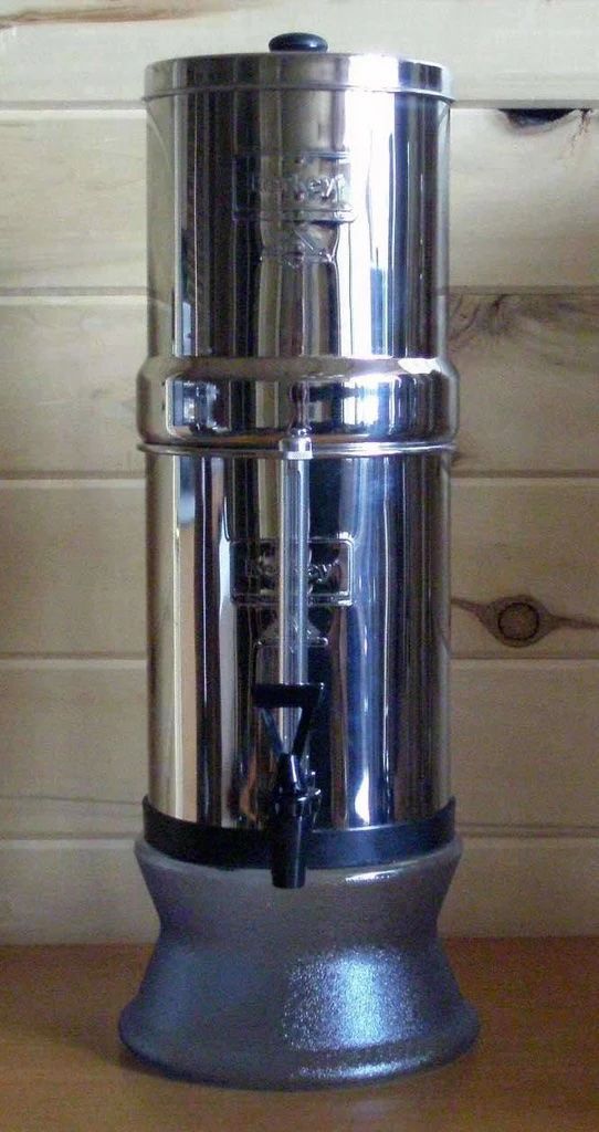 Stainless steel water filter with a spigot, on a glass base, against wooden planks.