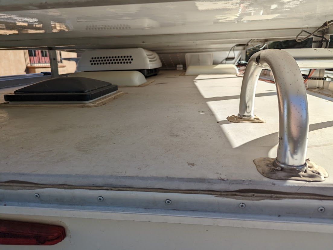 RV roof with AC unit, vent, grab bar, and sealant, shot outdoors.