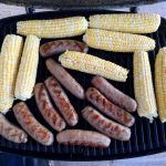 Corn on the cob and sausages are cooking on a grill.