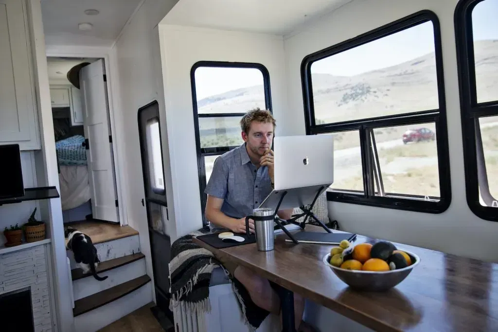 Man working on laptop inside a camper, near a window with a mountain view. A cat sits nearby.
