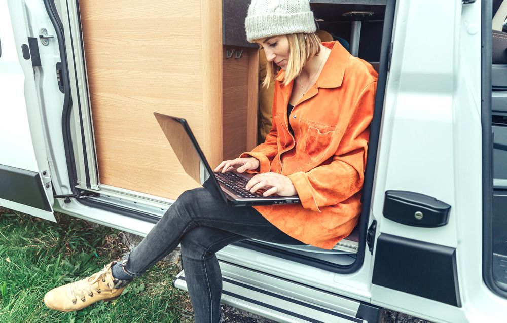Woman in orange shirt and beanie working on laptop in van doorway.