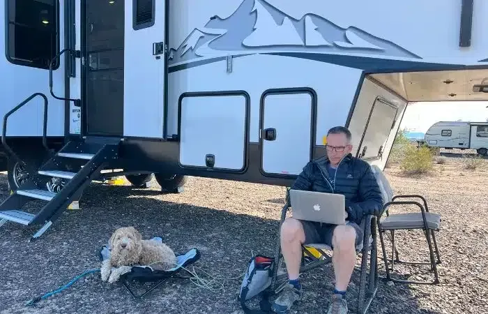 Man in chair using laptop outside RV with dog; desert setting.
