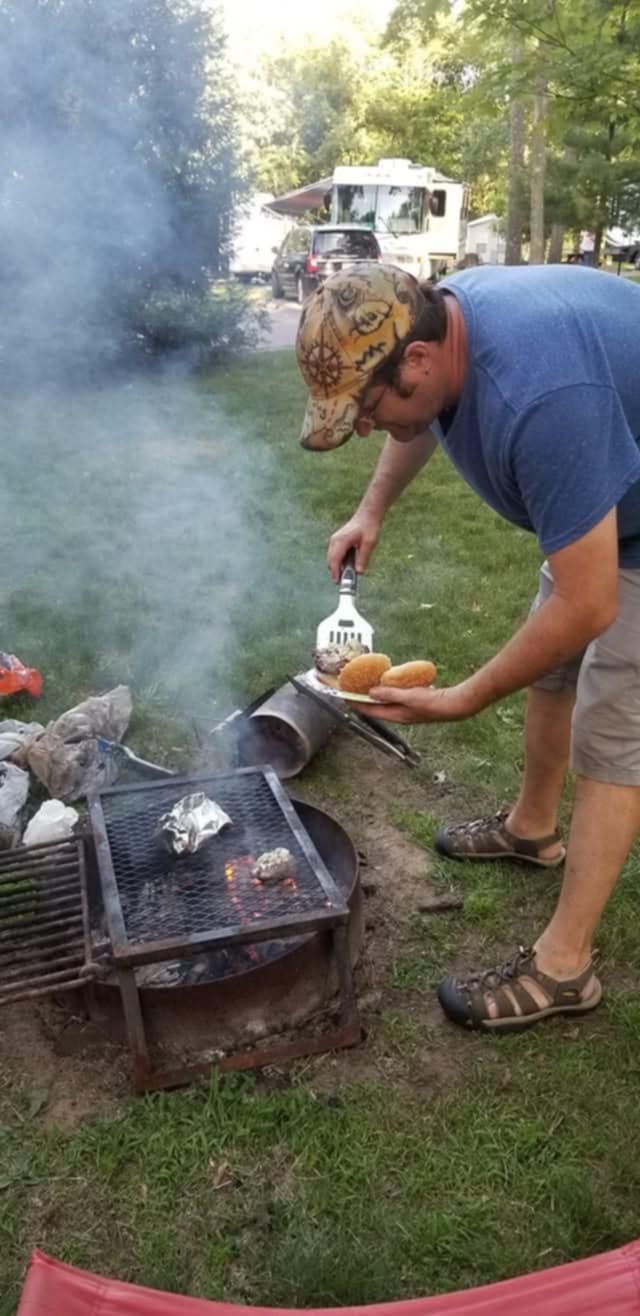 Man grilling food at a campsite, holding hot dog buns. A grill is over a fire pit, and smoke is rising.