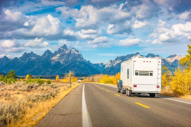 RV driving on road with mountains in background. Blue sky, fall foliage, and sunny.