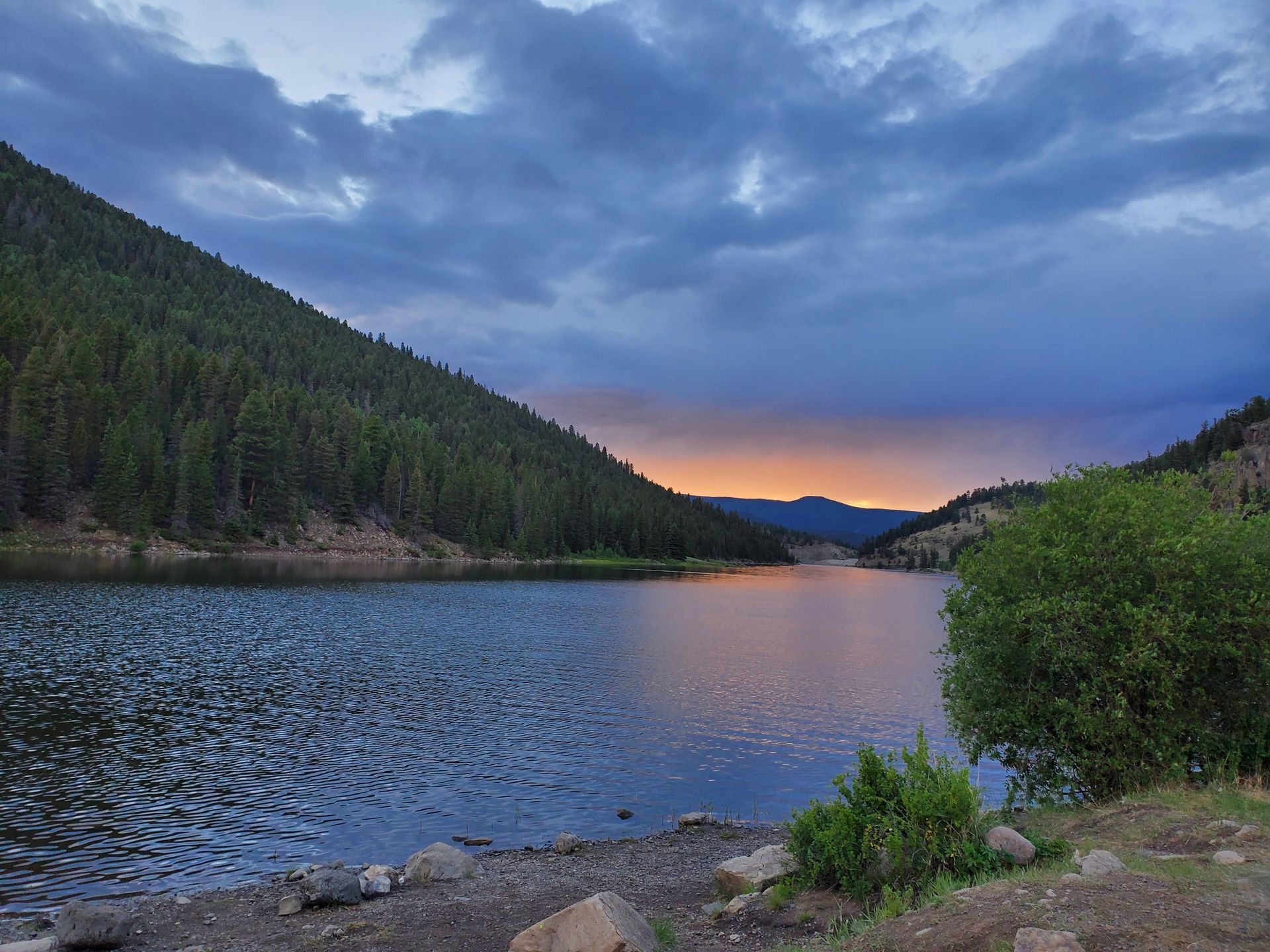 A lake surrounded by mountains and trees with a sunset in the background.