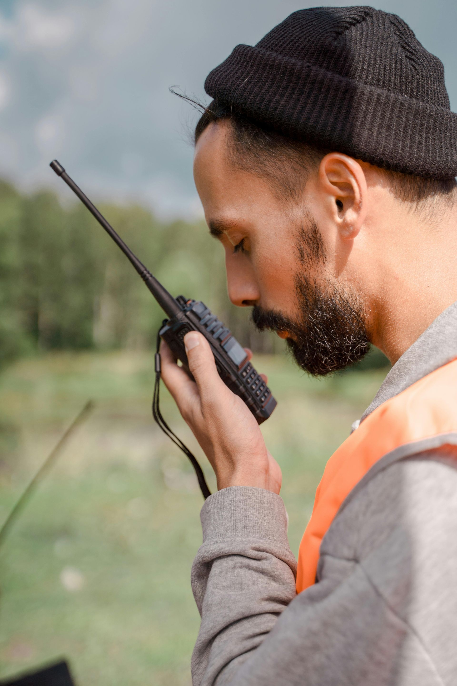 Man with a beard holding a walkie-talkie, wearing a beanie and orange vest, outdoors near trees.