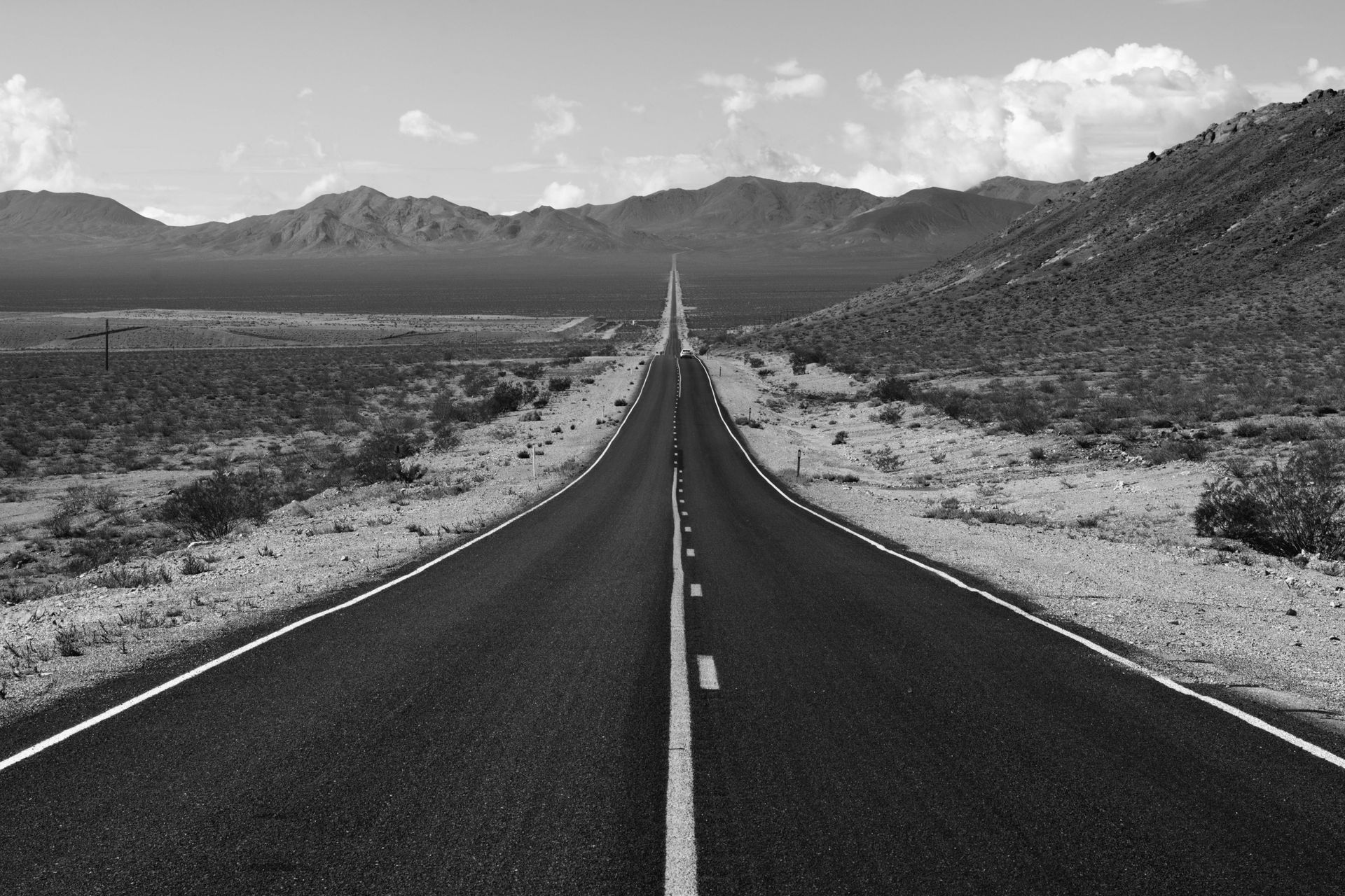Black and white photo of a long, straight road stretching into the distance, with mountains on the horizon.