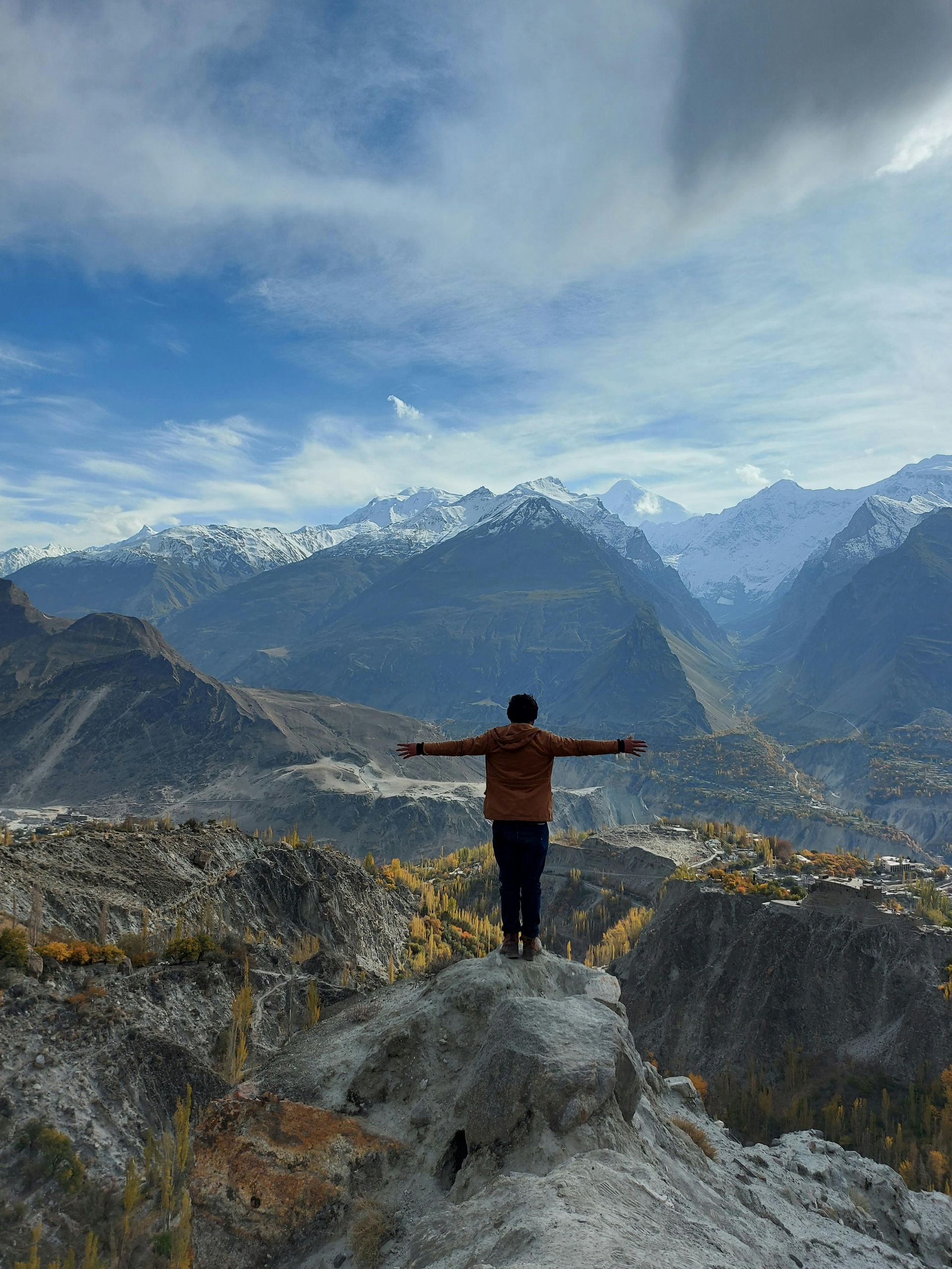 Person with arms outstretched atop a mountain overlooking a valley with snowy peaks, under a cloudy blue sky.