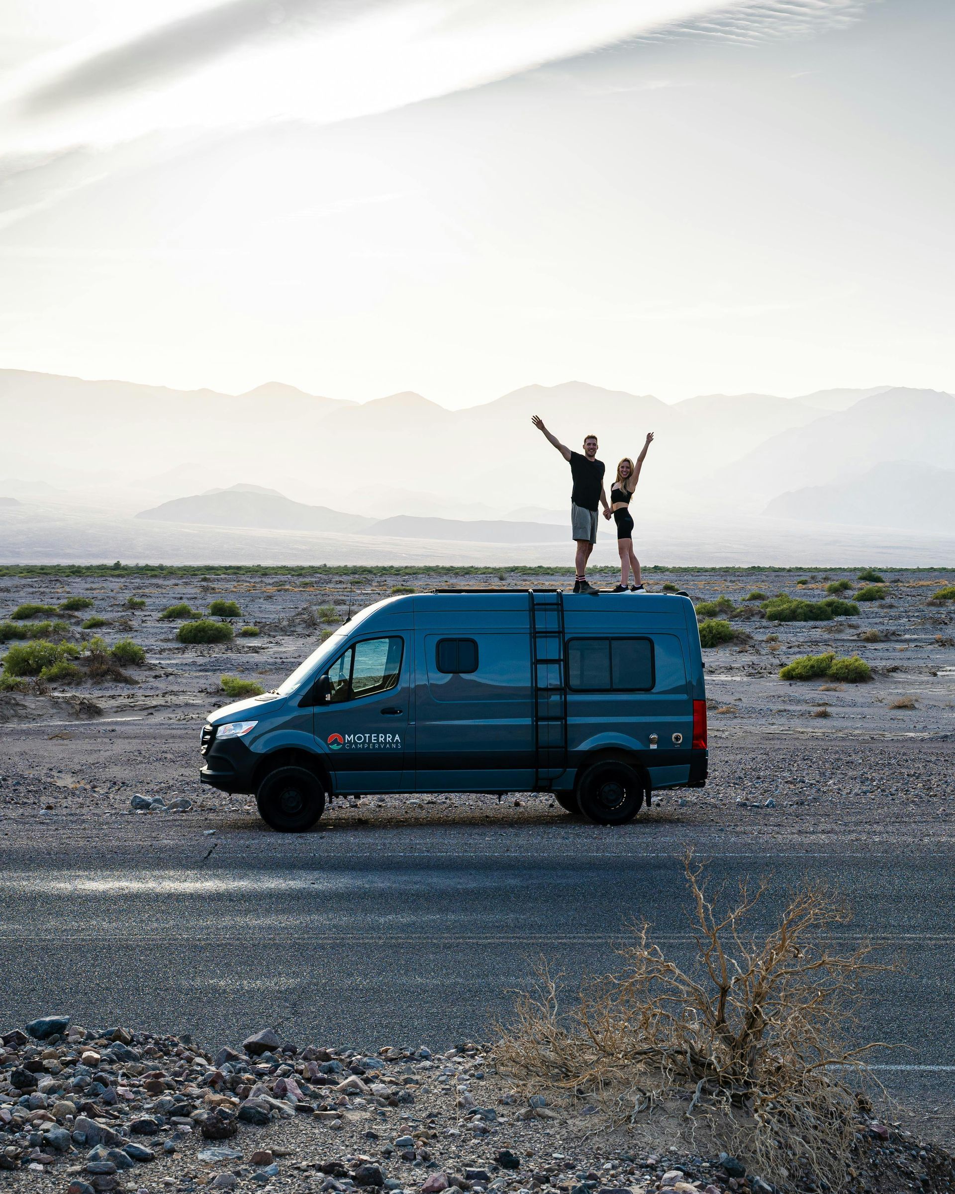 Couple standing on the roof of a blue van, arms raised, in a desert landscape.