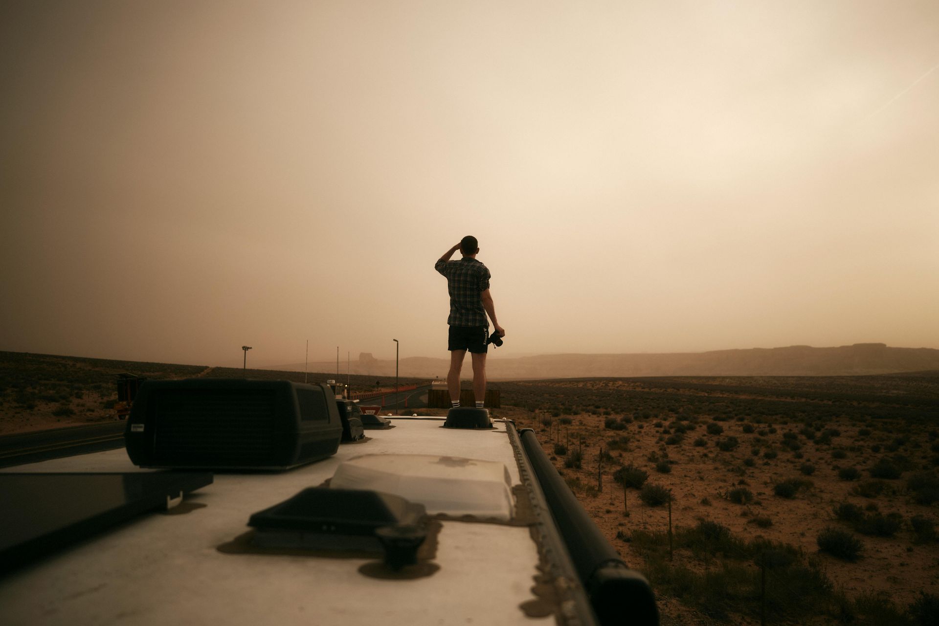 Person standing on the roof of a vehicle in a desert at sunset, looking into the distance.