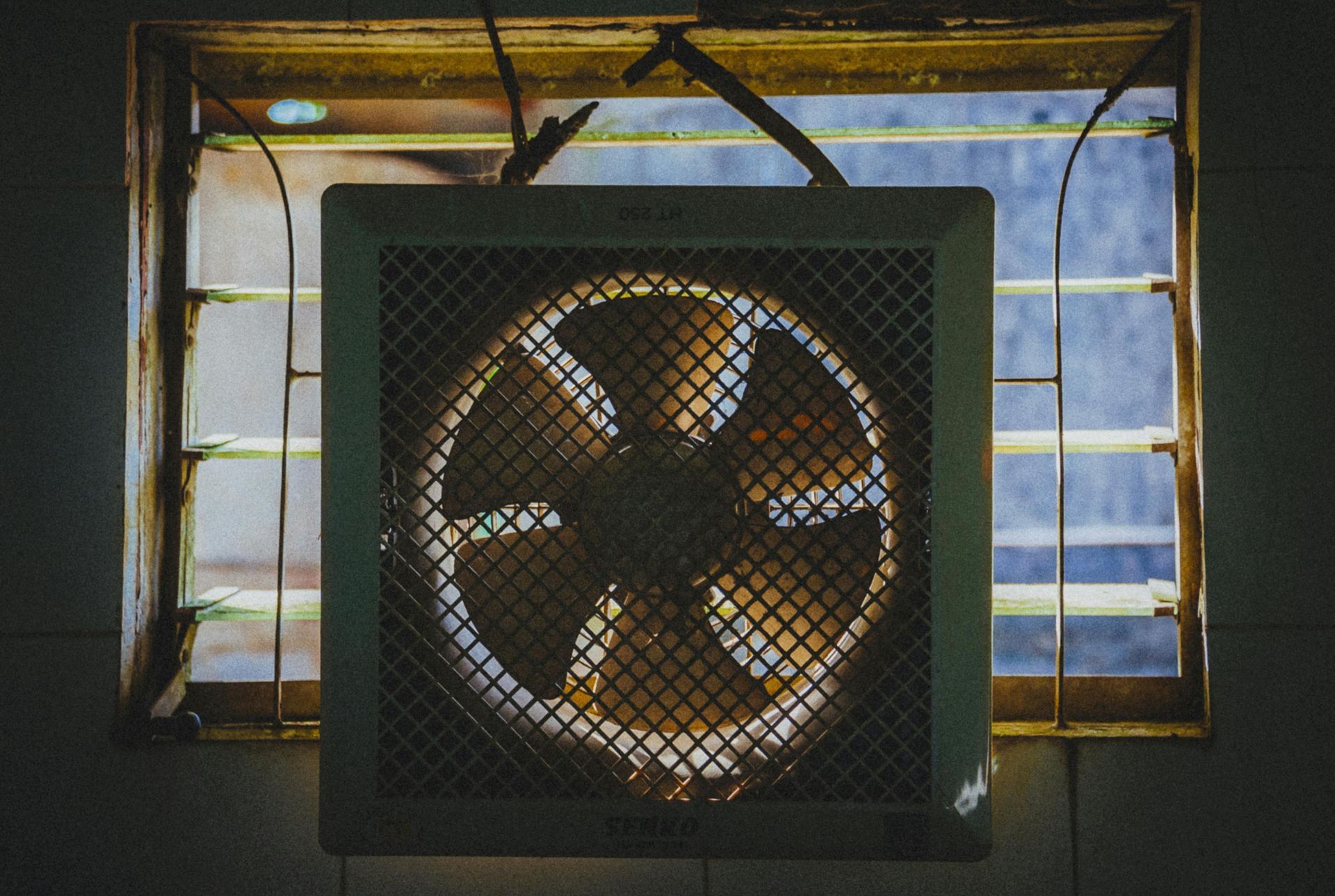 Square fan with protective metal grill in front of a window. Blades visible through the fan's grill.