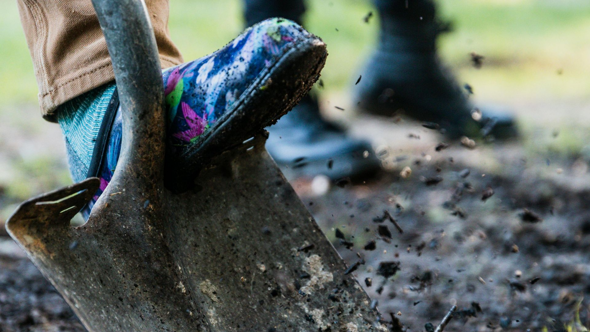 Foot in patterned boot presses shovel into dark soil; another person in boots in background.