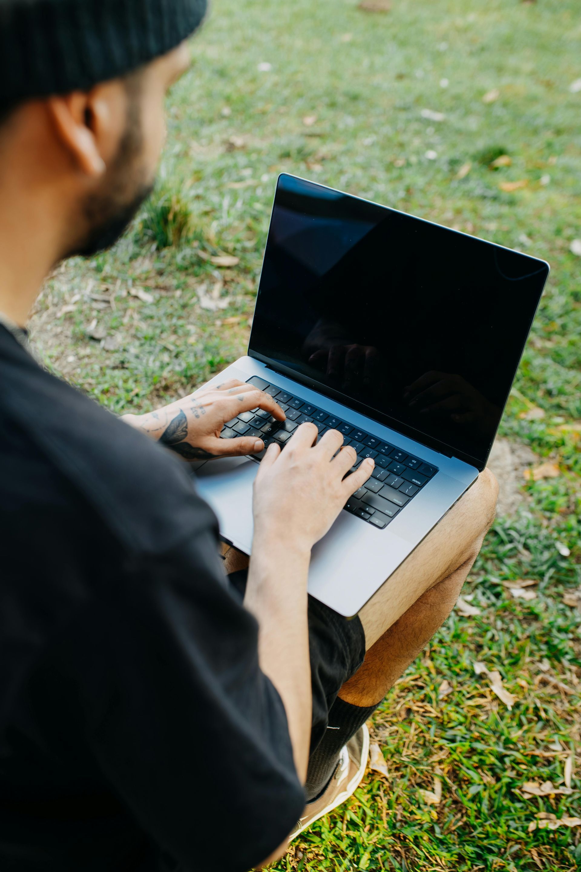 Person using a laptop outdoors, typing on the keyboard. Black t-shirt, beanie, and tattoo. Green grass background.