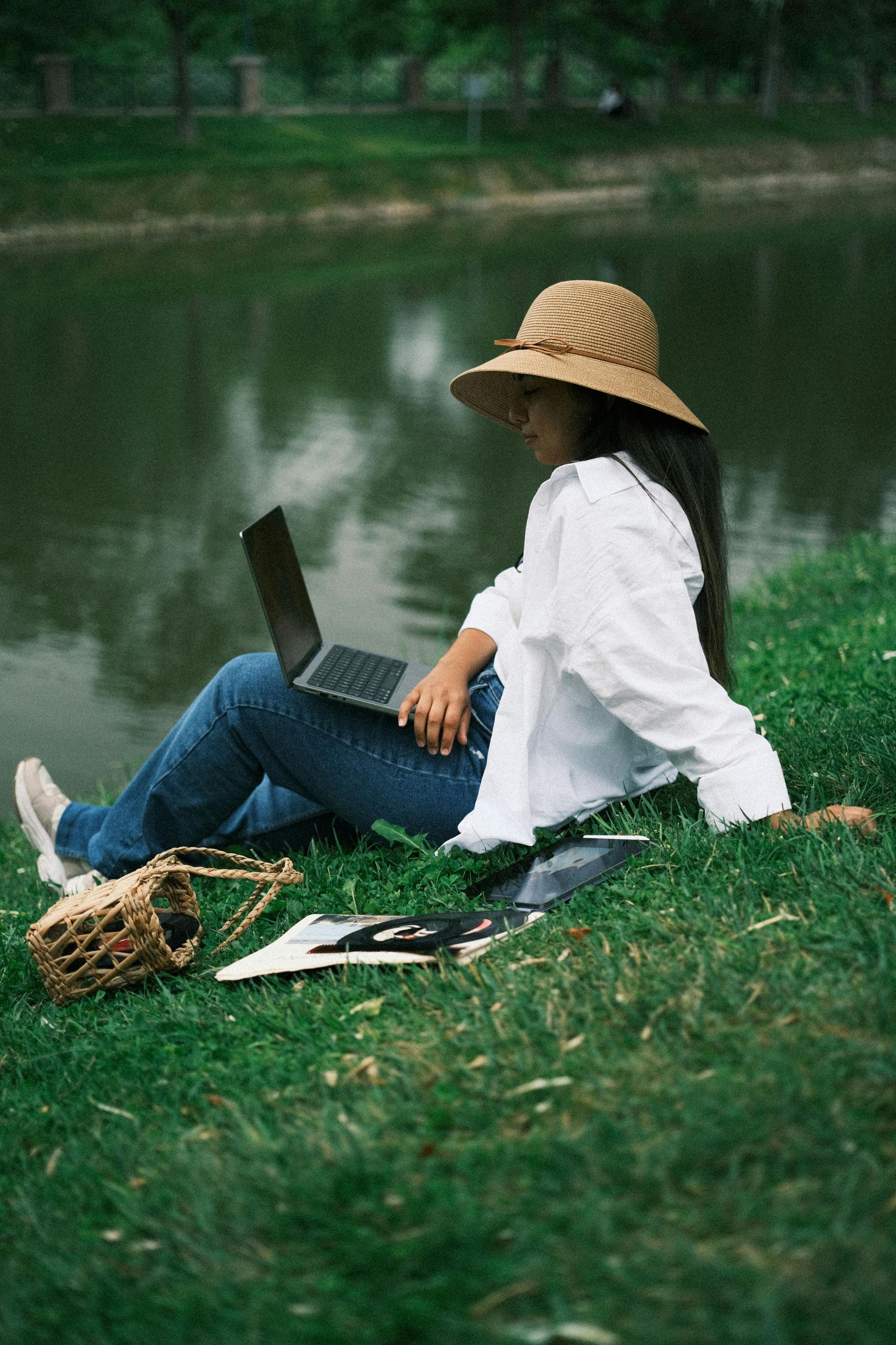 Person wearing a hat and using a laptop on the grass near water. A basket and notebook are nearby.