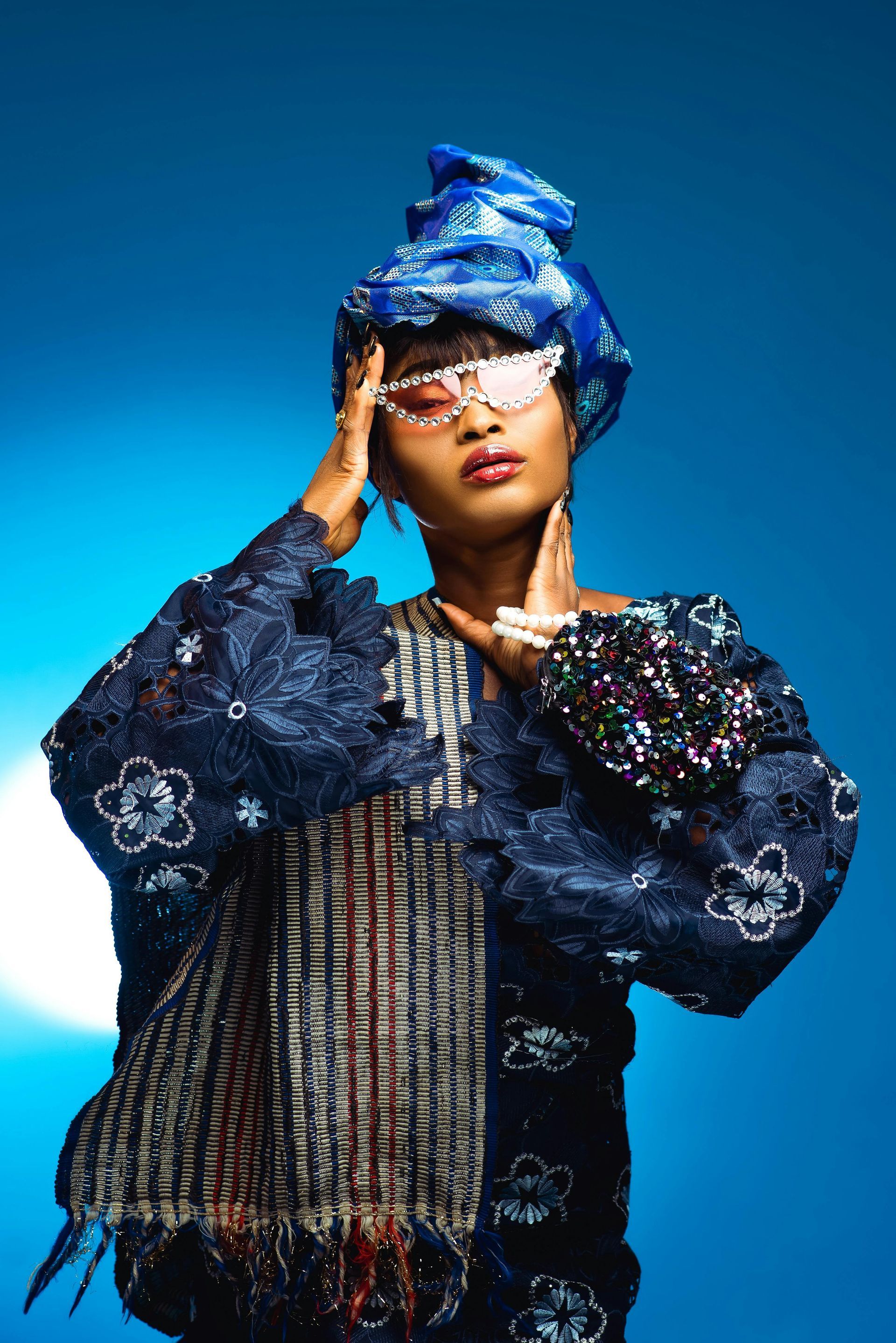 Woman in blue ornate outfit and head wrap, wearing bedazzled sunglasses, hands near face, blue backdrop.