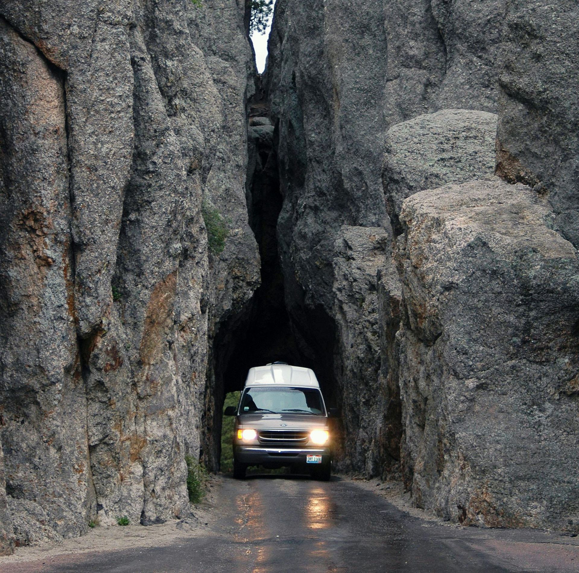 Van driving through a narrow tunnel carved through rock formations in a mountainous region.