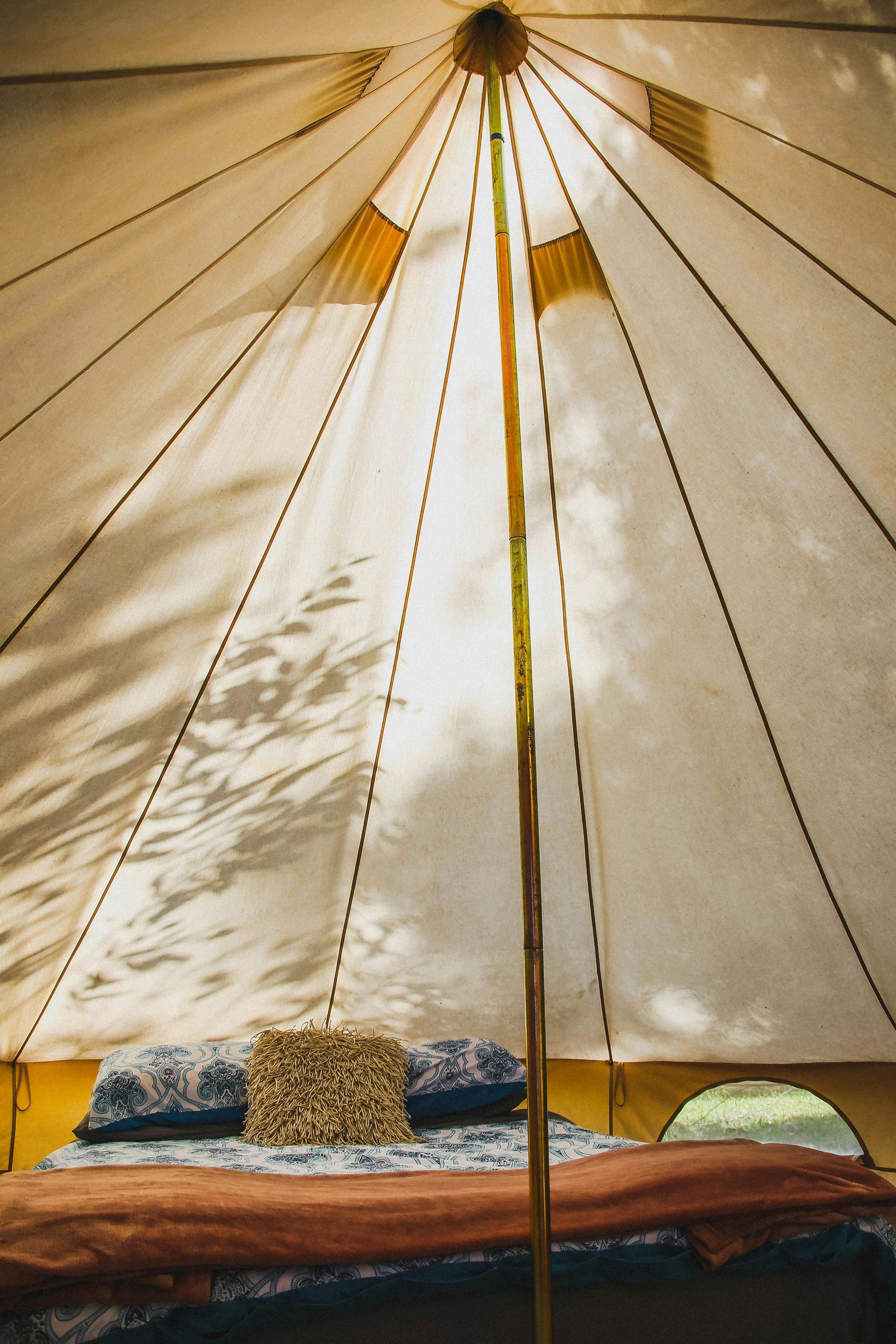 Interior of a canvas bell tent, furnished with a bed, pillows, and blanket; light streams in.