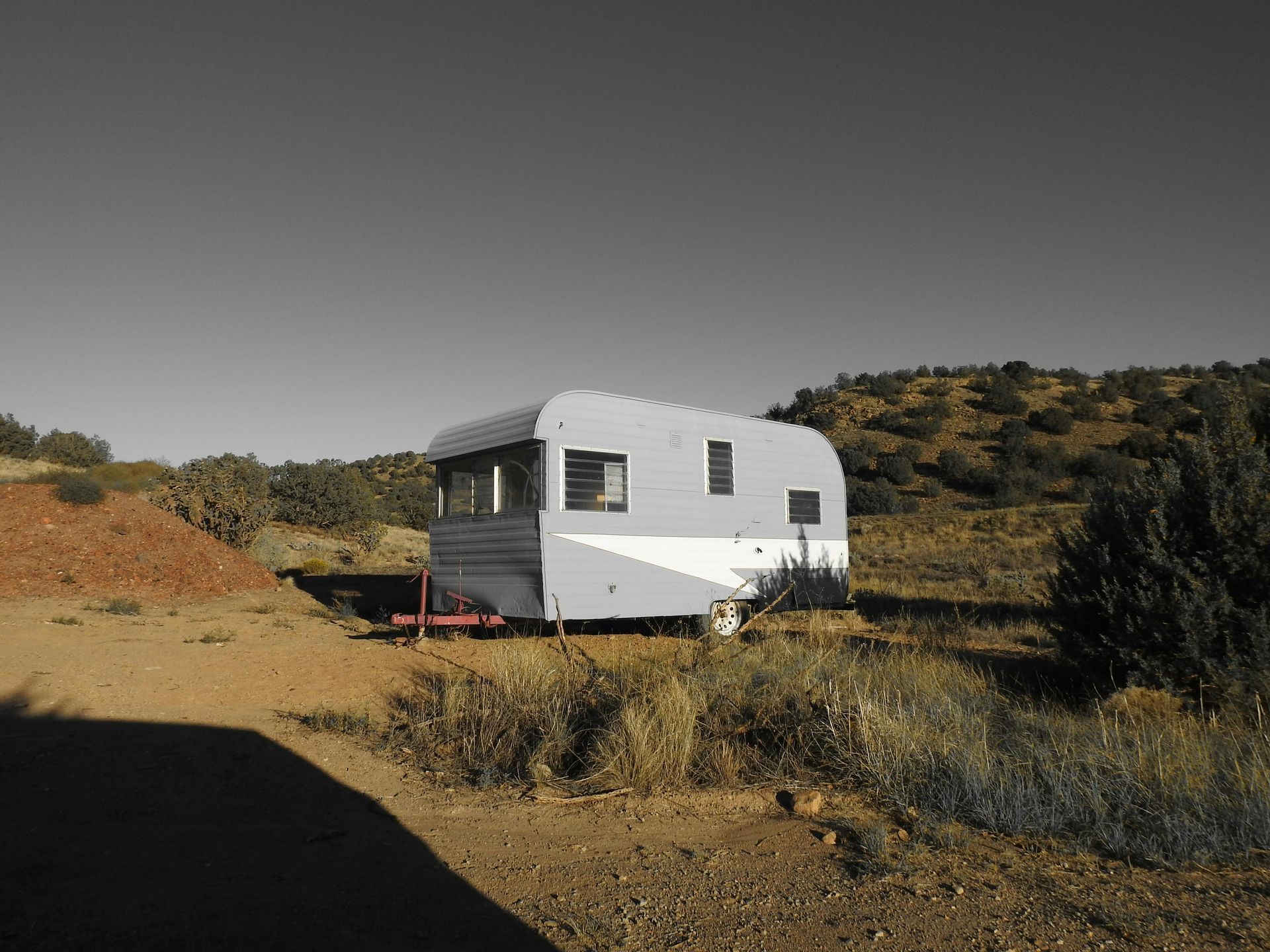 Gray and white vintage travel trailer parked in a dry, grassy field with shrubs and a hill in the background.