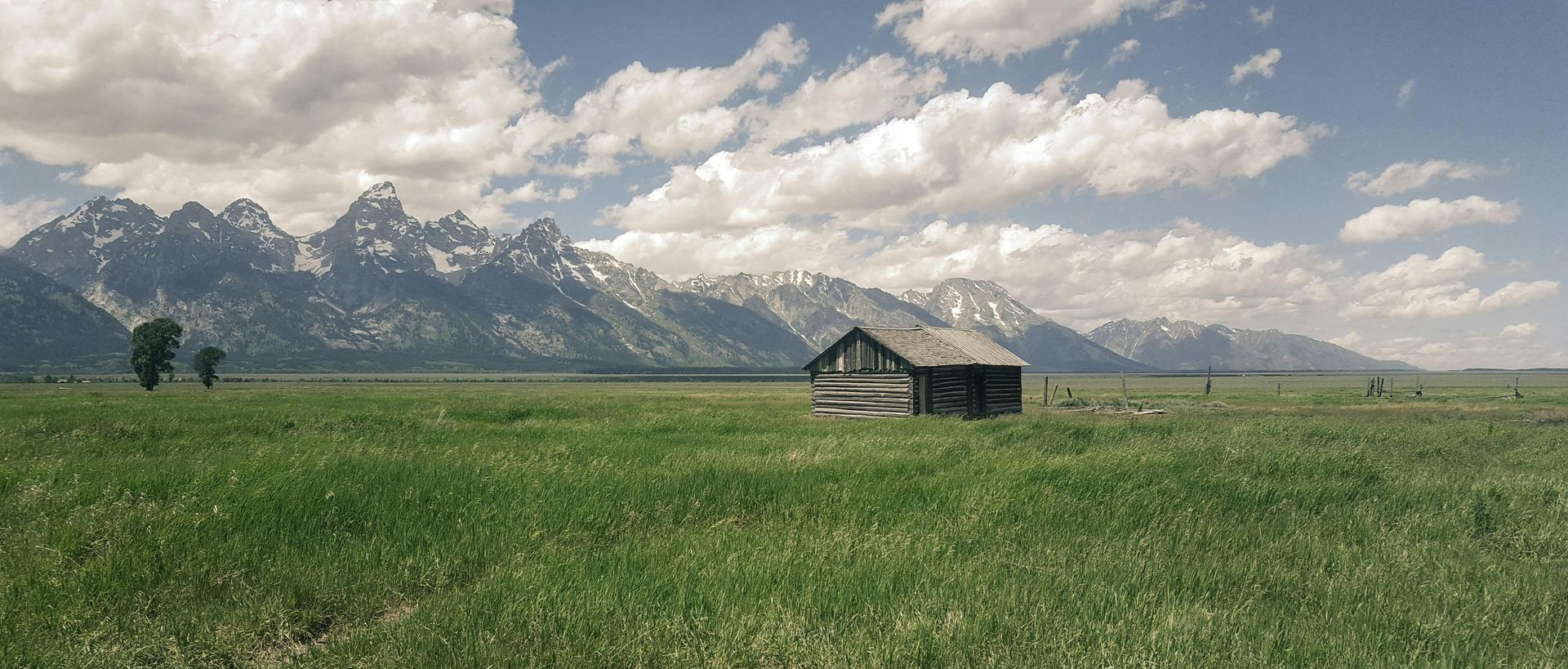 A grassy field with a small wooden cabin, mountains in the background, under a cloudy sky.