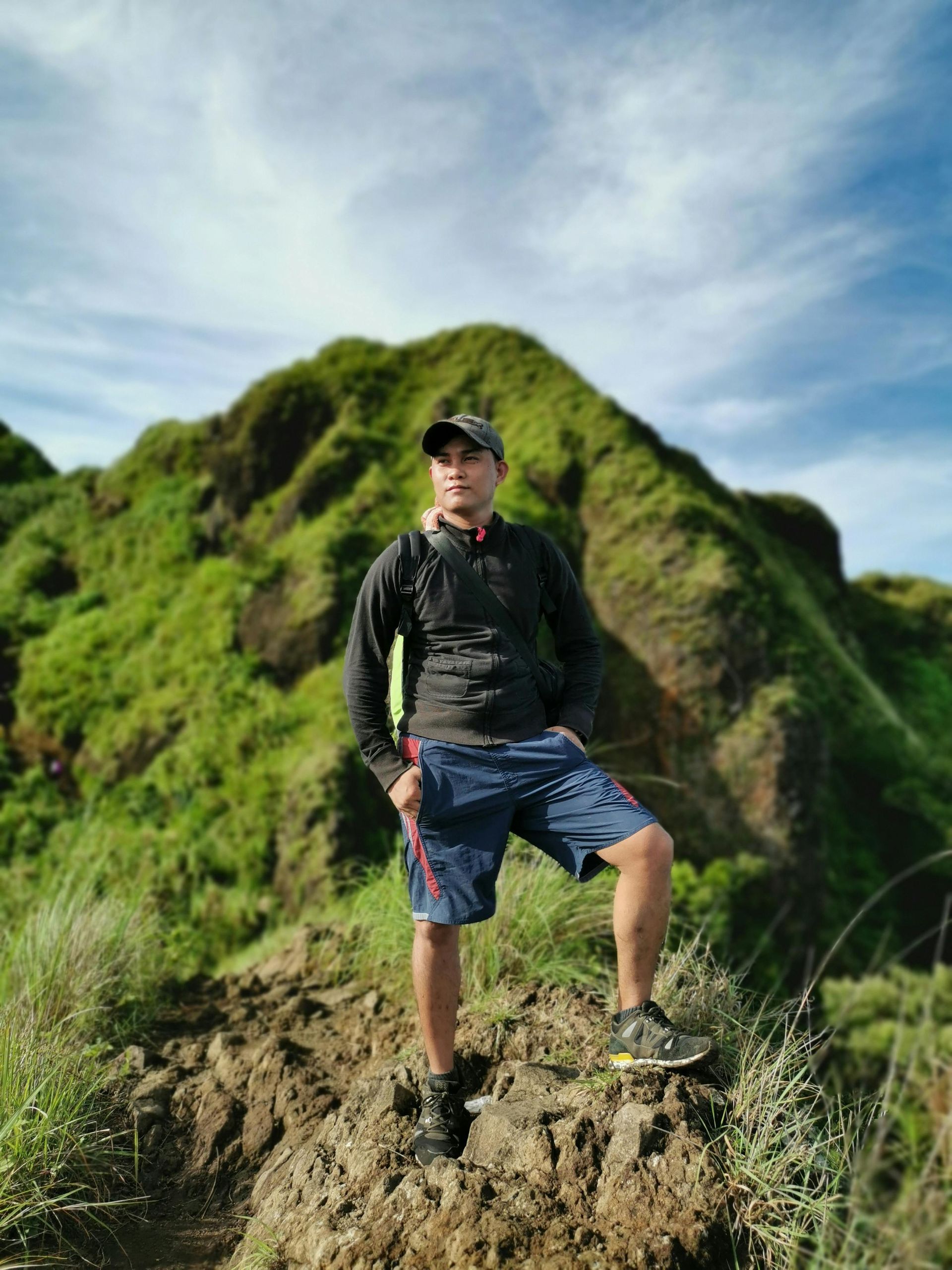 Man standing on a hillside, looking toward the side, wearing a hat, shorts, and backpack. Green hills and blue sky background.