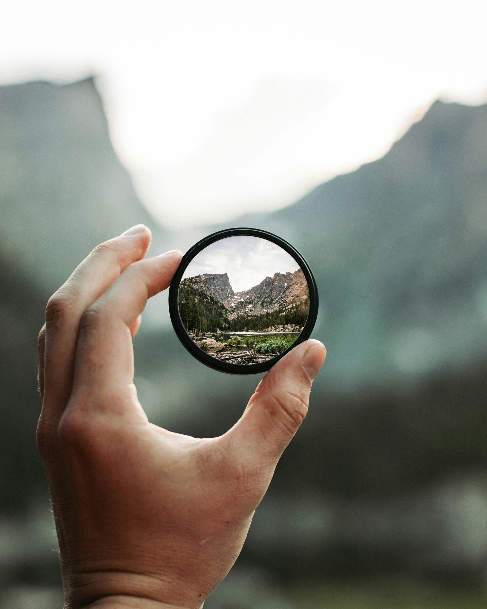 Hand holding a circular lens, framing a mountain landscape with trees and a body of water.