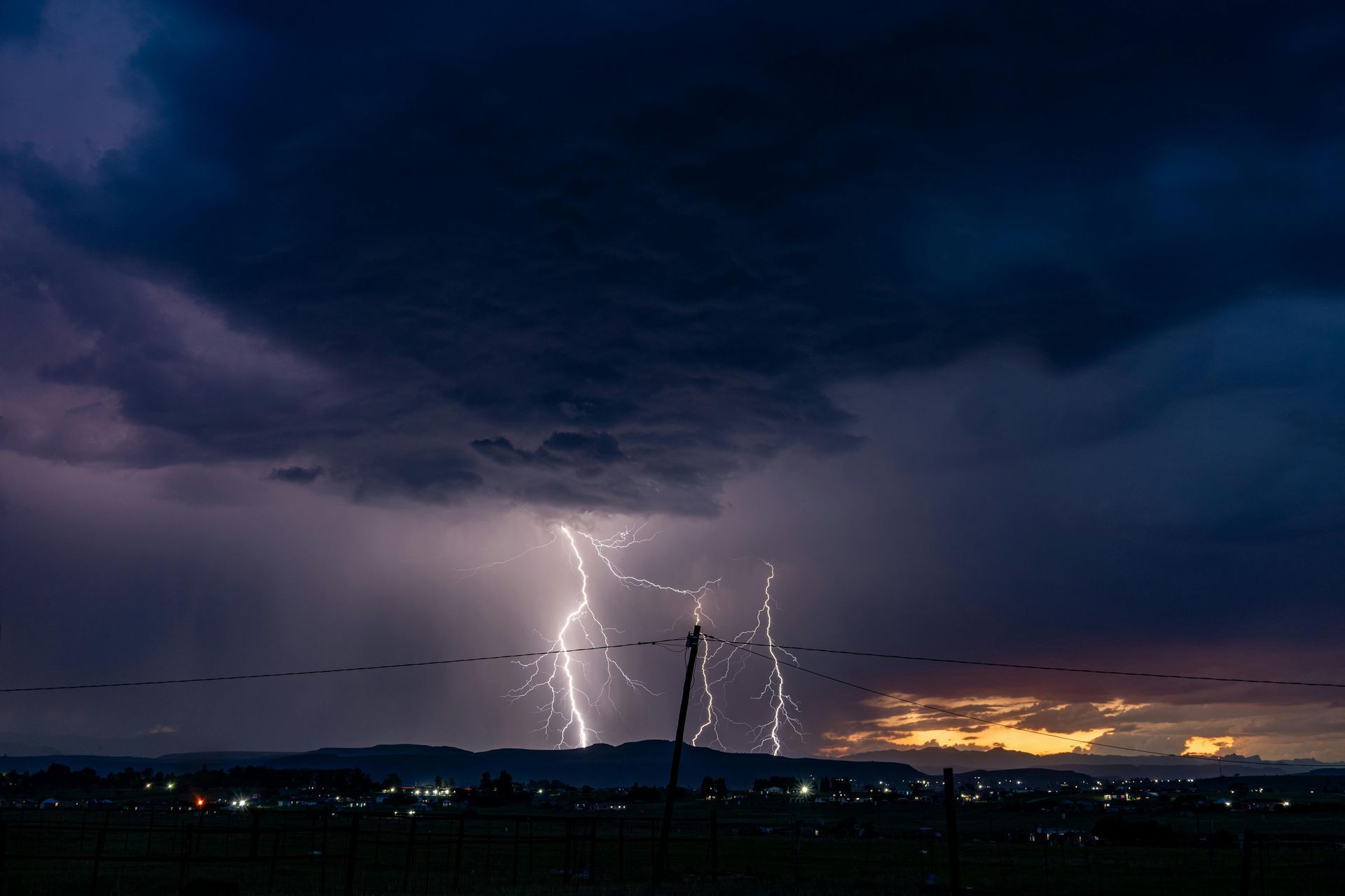 Lightning bolts over a dark stormy landscape at dusk, with distant city lights and a glowing horizon