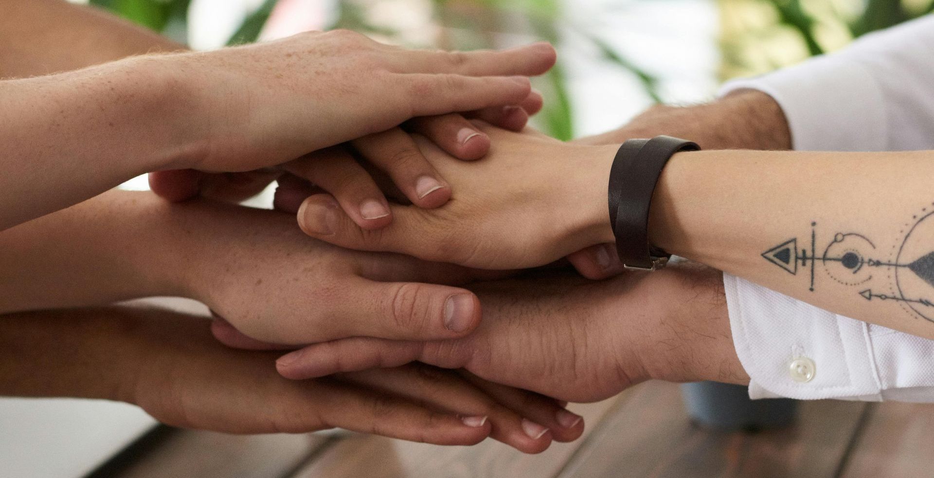 Hands stacked in a pile, demonstrating teamwork and collaboration. One arm has a tattoo.