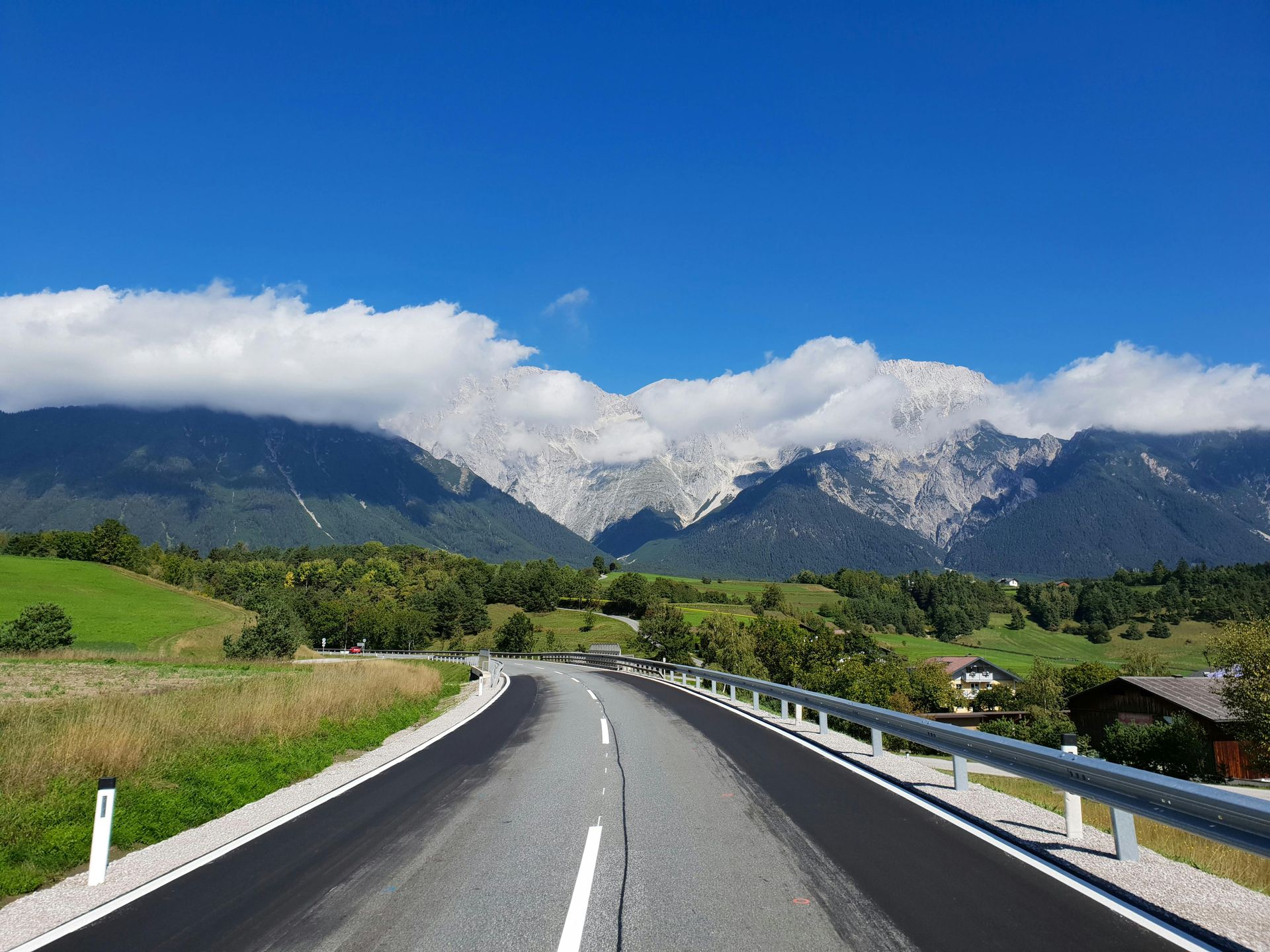 Road leading toward snow-capped mountains under a blue sky, with green fields and trees on either side.