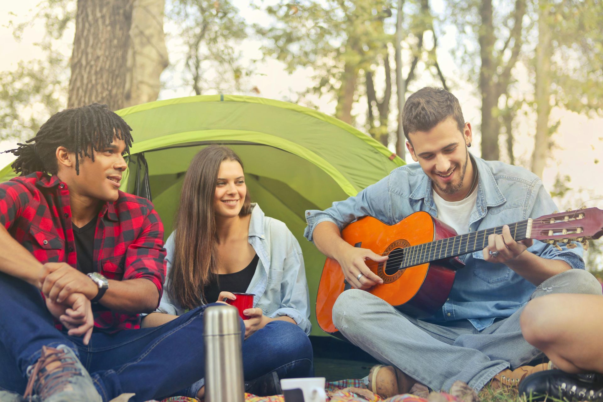 Three people camping, one plays guitar. Tent and trees in background.