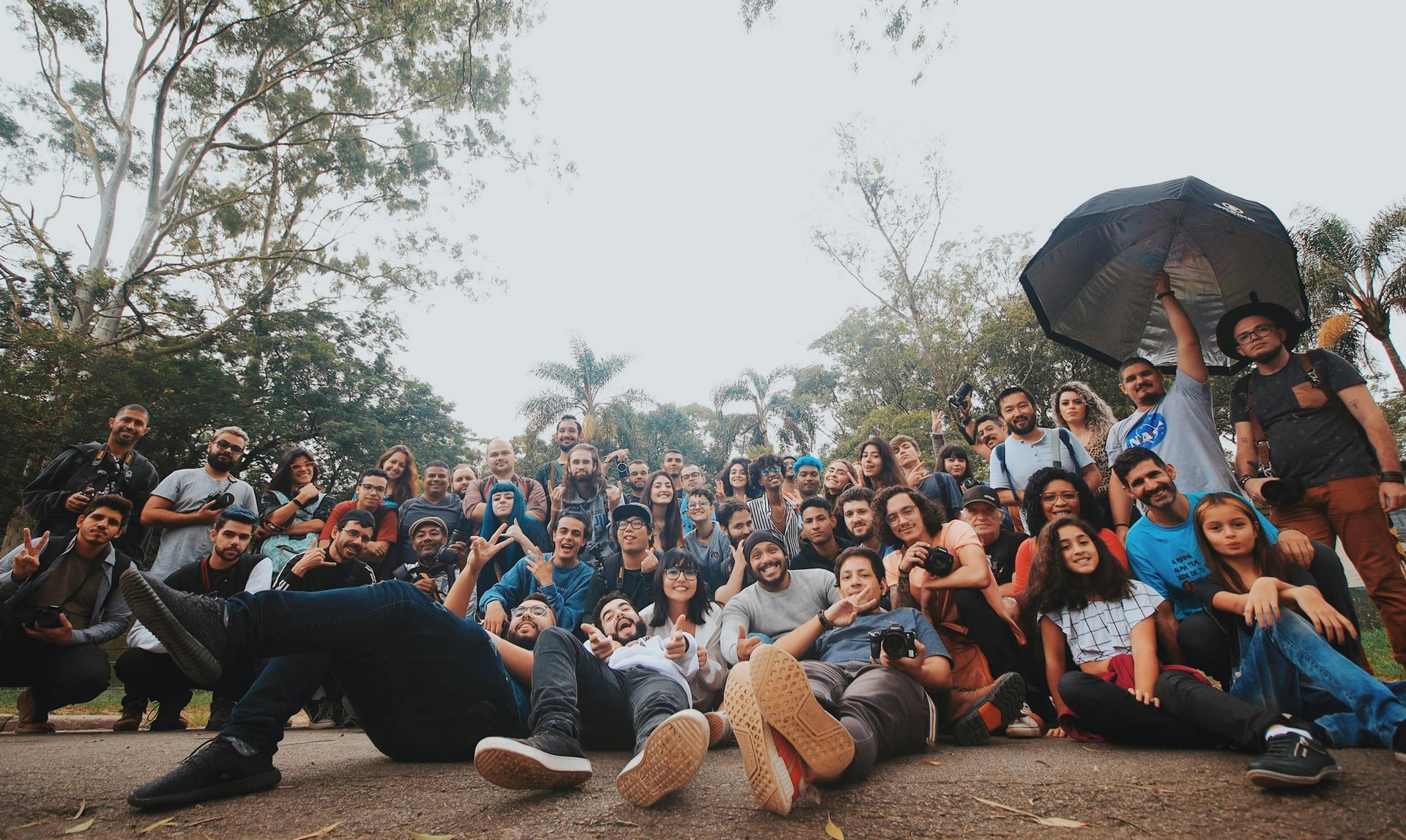 Large group of people gathered outdoors, some seated, some standing, smiling. Overcast sky, trees in background.