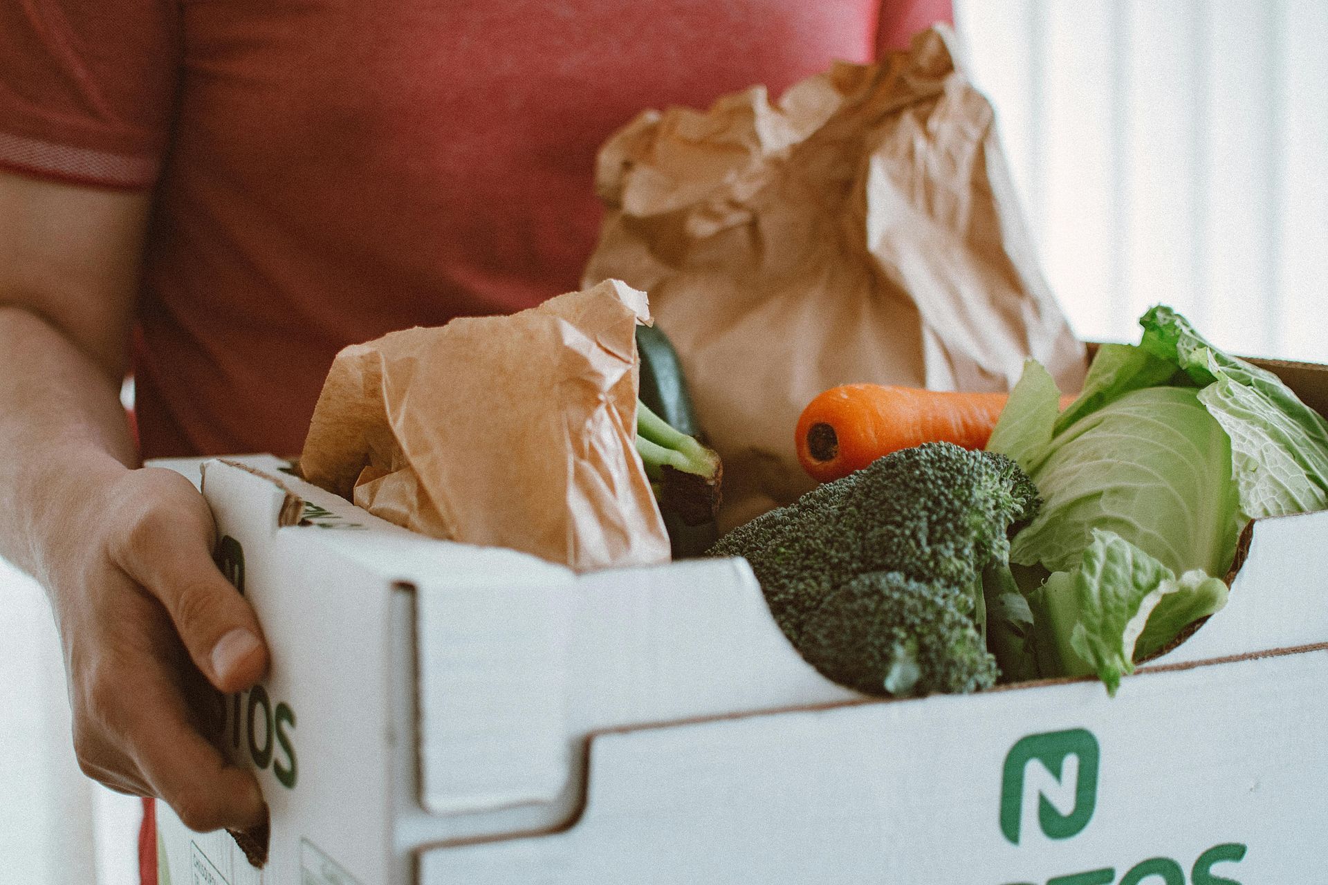 Person holding a box of groceries including vegetables and a paper bag.