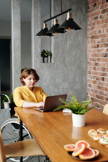 Woman in yellow shirt typing on laptop at wooden table, near brick wall and plant.