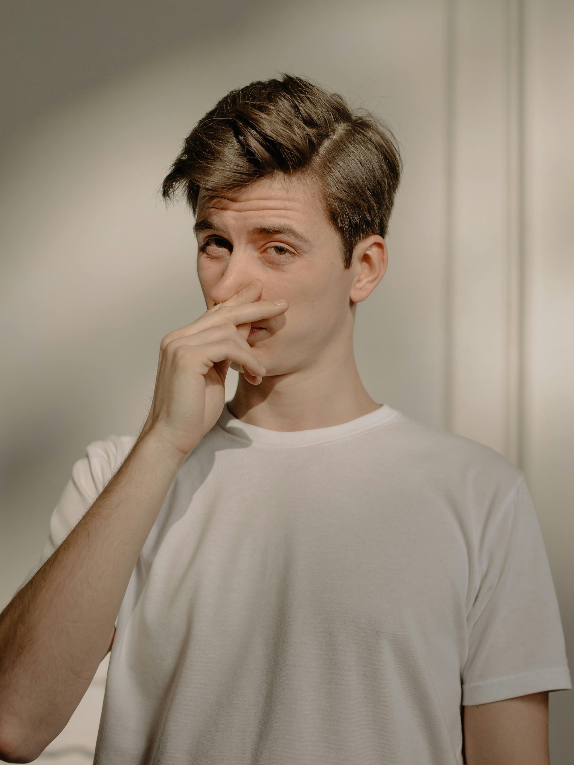 Person in a white T-shirt pinching their nose while standing indoors by a beige wall.