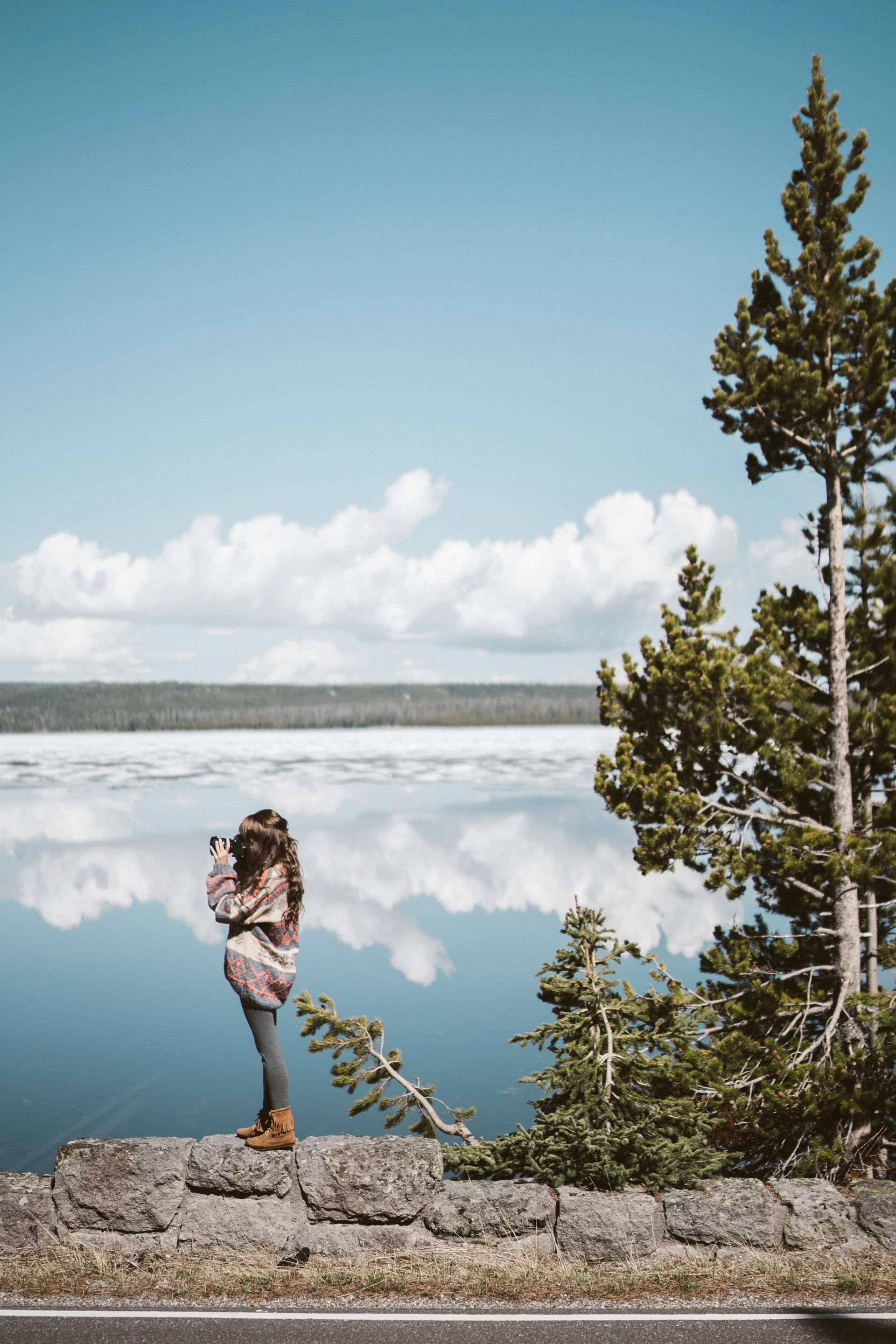 Woman photographing lake and sky from a stone wall, with pine trees.